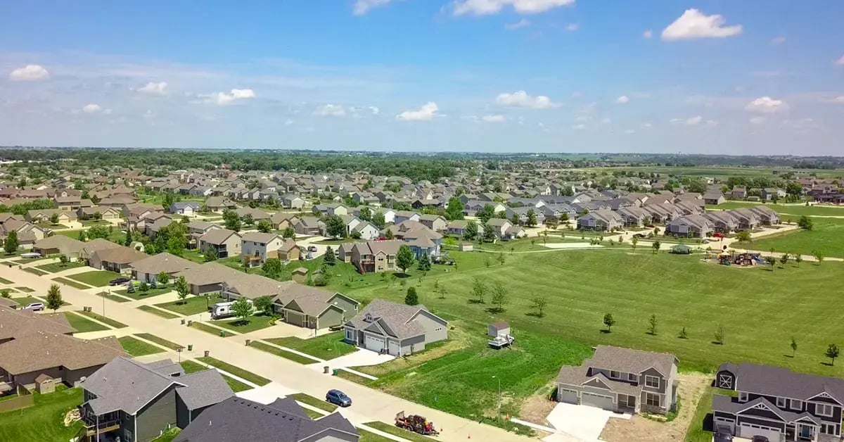 Aerial view of a large suburban neighborhood in Altoona, Iowa, featuring rows of single-family homes, wide streets, green lawns, and an open park area under a bright blue sky with scattered clouds.