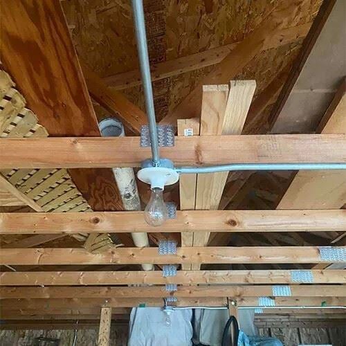 Exposed light bulb fixture mounted on unfinished ceiling joists with visible electrical conduit in a basement or attic space.