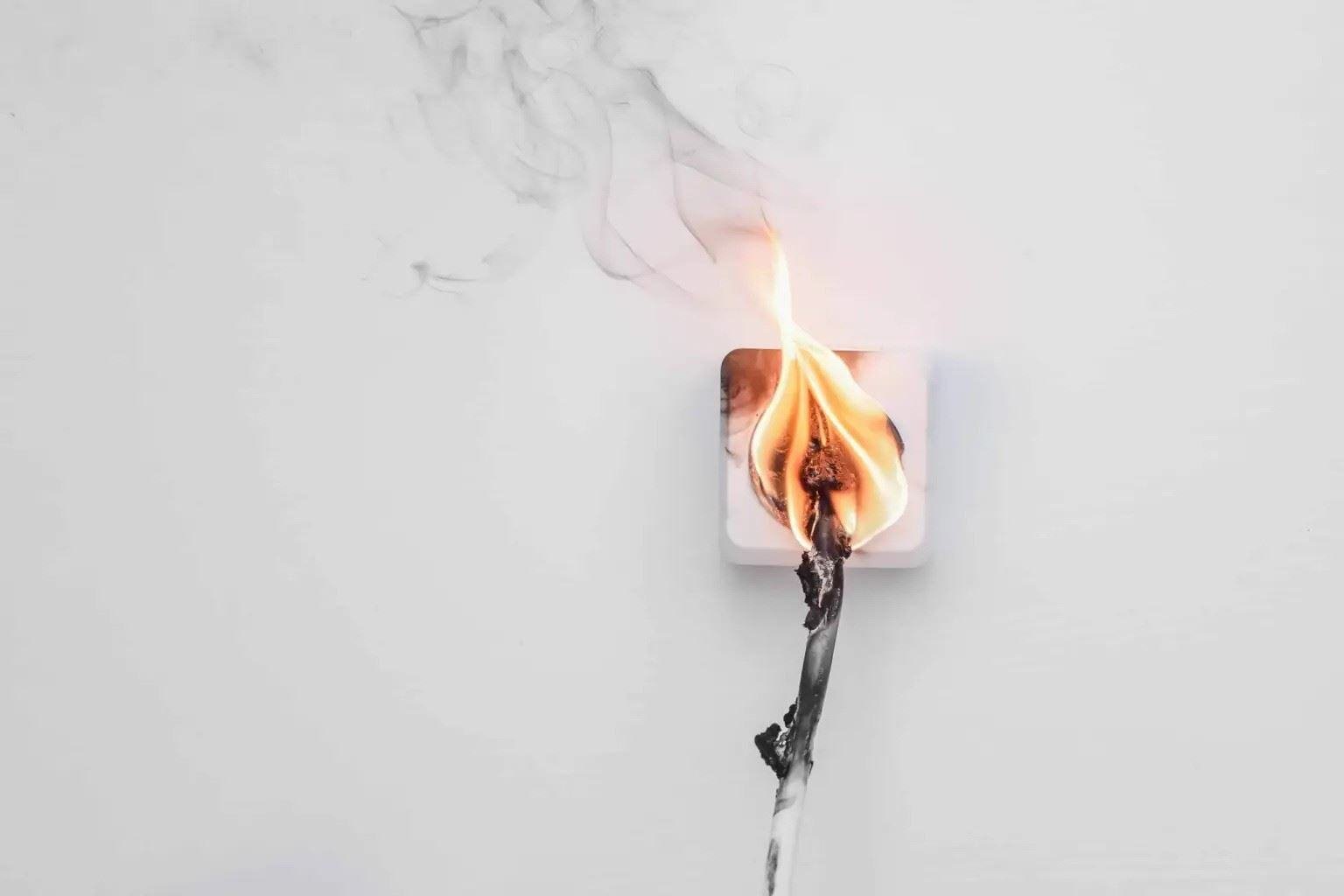 Electrical outlet with a burning plug and visible flames on the cord against a white wall.