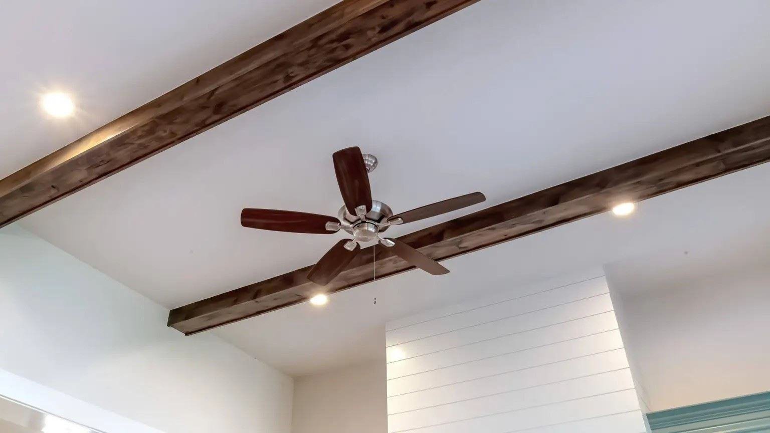 Ceiling fan with wooden blades and recessed lighting installed between exposed ceiling beams in a modern living space.
