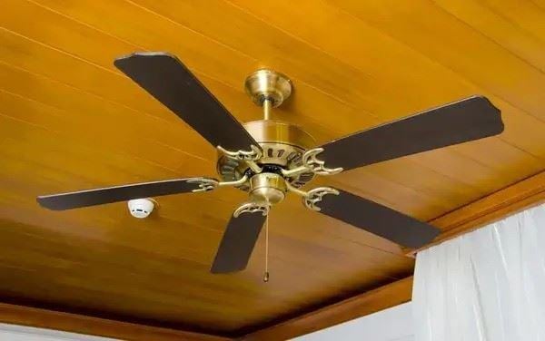 “Ceiling fan with brass housing and dark wooden blades mounted on a wood-paneled ceiling next to a smoke detector