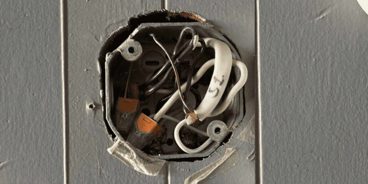 An open electrical junction box recessed into a gray paneled wall, showing a cluster of black, white, and bare copper wires. Several connections are secured with bright orange WAGO lever-nut connectors, which are clearly visible in the lower left portion of the box. The surrounding wall paint is chipped, and a piece of frayed white tape is partially attached to the metal box. 