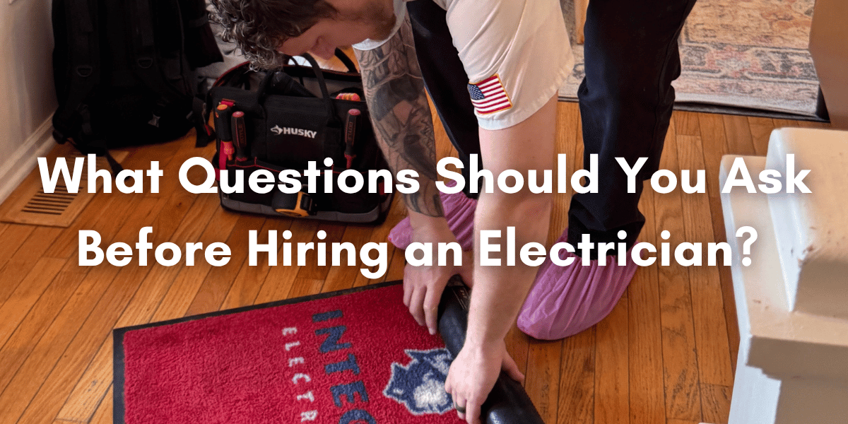 An electrician kneels on a hardwood floor while unrolling a red Integra Electrical floor mat beside a tool bag, with overlaid text reading, “What Questions Should You Ask Before Hiring an Electrician?”