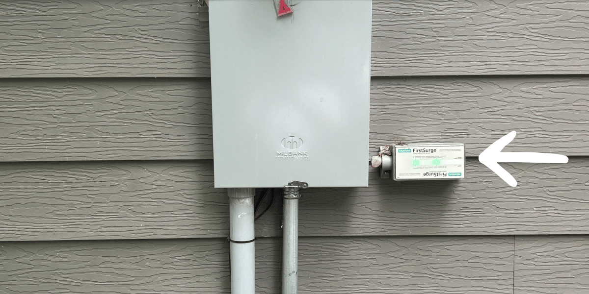Whole-house surge protector mounted beside an outdoor electrical panel, highlighted by a white arrow.